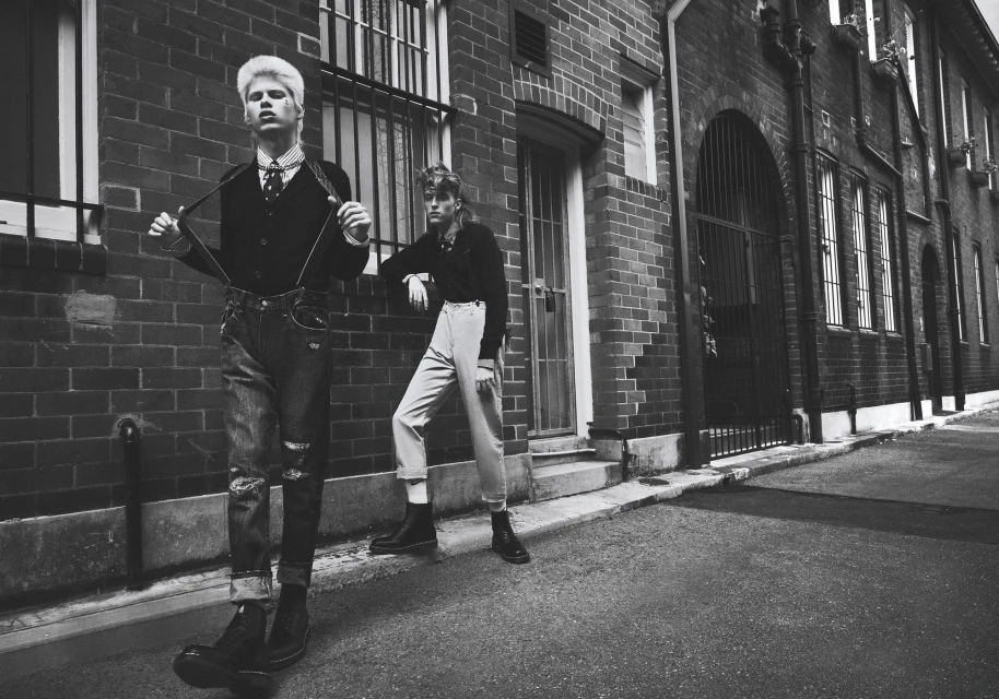 Two young adult fashion models in black and white stand outside a brick building in jeans, suspenders, and boots in an outdoor setting.