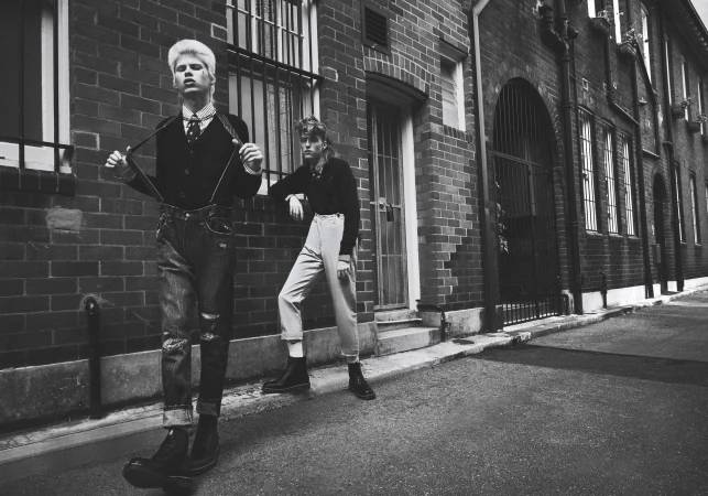 Two young adult fashion models in black and white stand outside a brick building in jeans, suspenders, and boots in an outdoor setting.