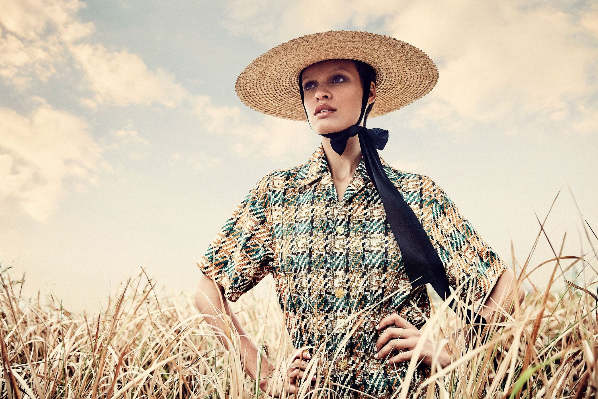Woman in straw hat and patterned shirt posing in field of tall grass with a cloudy sky. Fashion editorial style outdoor shot.