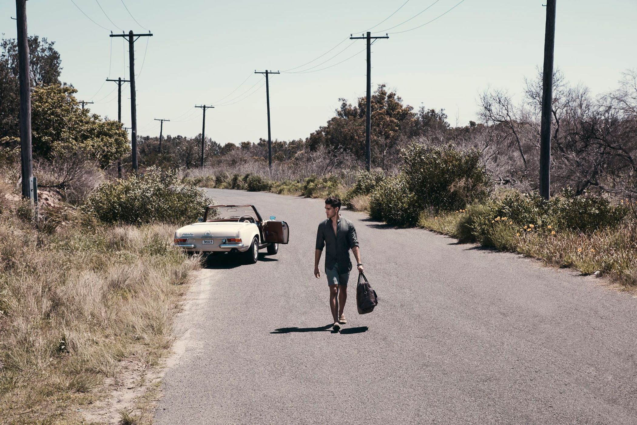 Adult male walks on a rural road carrying a duffel bag with a vintage convertible car parked on the side of the road.