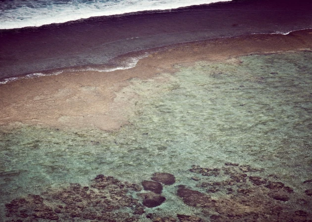 Aerial view of ocean water meeting the shore. Waves are breaking in the distance, with varying depths and textures of water visible.