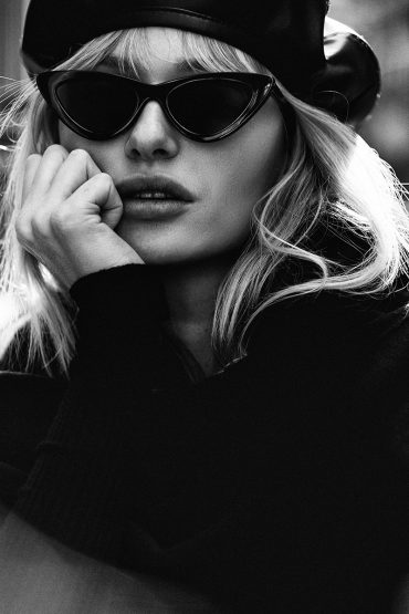 Monochrome portrait of a woman with blonde wavy hair wearing dark sunglasses and a black beret posing indoors.