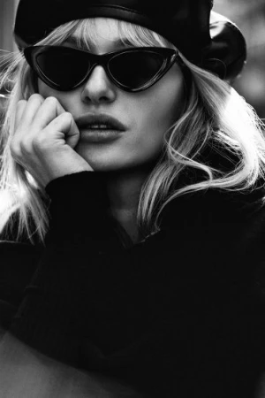 Close-up black and white portrait of a blonde woman wearing cat-eye sunglasses and a beret, with her hand resting against her chin.