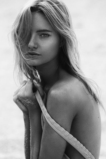 Black and white portrait of a blonde woman with her hands to her chest on an overcast beach with soft, diffused natural light.