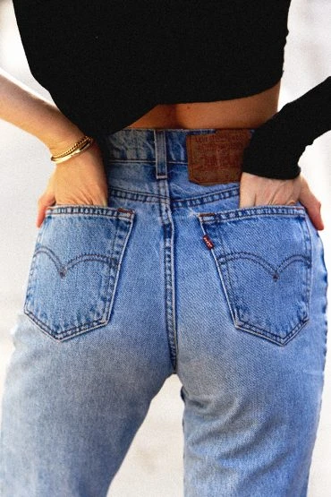 Back view of an adult female wearing light-wash blue jeans with her hands in her pockets, paired with a black long-sleeved top.