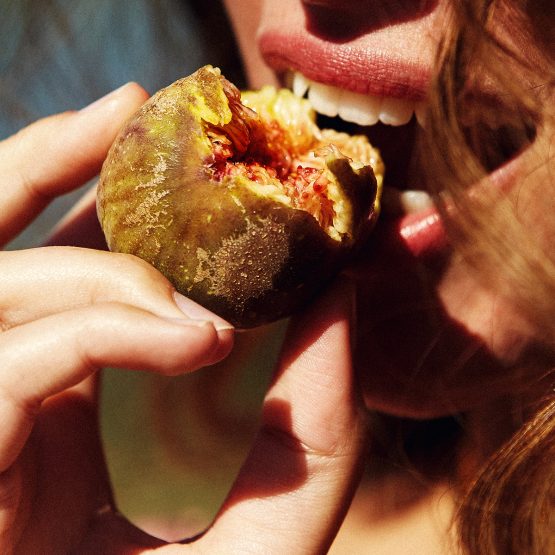 Close-up of a woman biting into a ripe fig, showcasing its texture and color against a blurred, natural background.