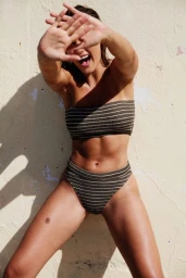 A smiling woman wearing a striped two-piece swimsuit holding her hands up in front of her face against a textured cream wall.