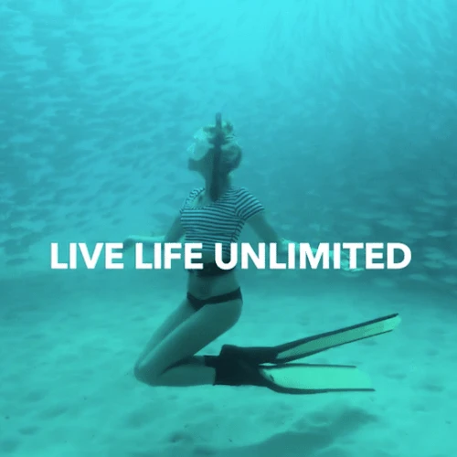 Woman underwater kneeling on the ocean floor wearing black and white striped shirt and black bikini, surrounded by schools of fish.