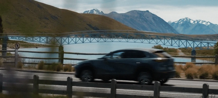 Dark-colored SUV drives along a road with a bridge over a body of water and mountains in the background under a bright sky.