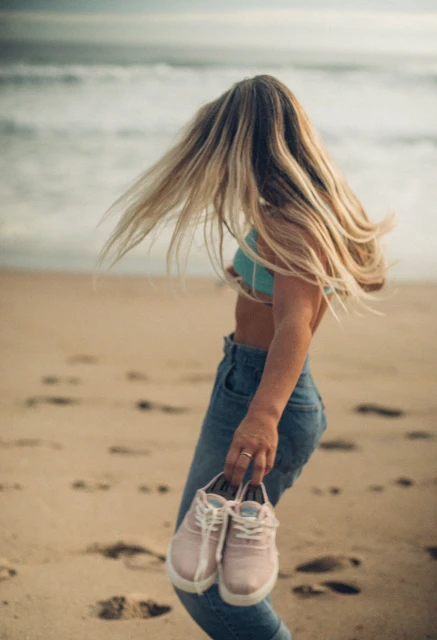 A woman with blonde hair in blue jeans and a bikini top stands on a sandy beach, holding a pair of pink shoes near ocean waves.