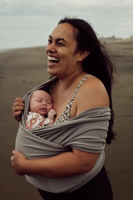 Happy mother carrying her newborn baby in a sling on a beach, both smiling in an eye-level shot with soft natural light.