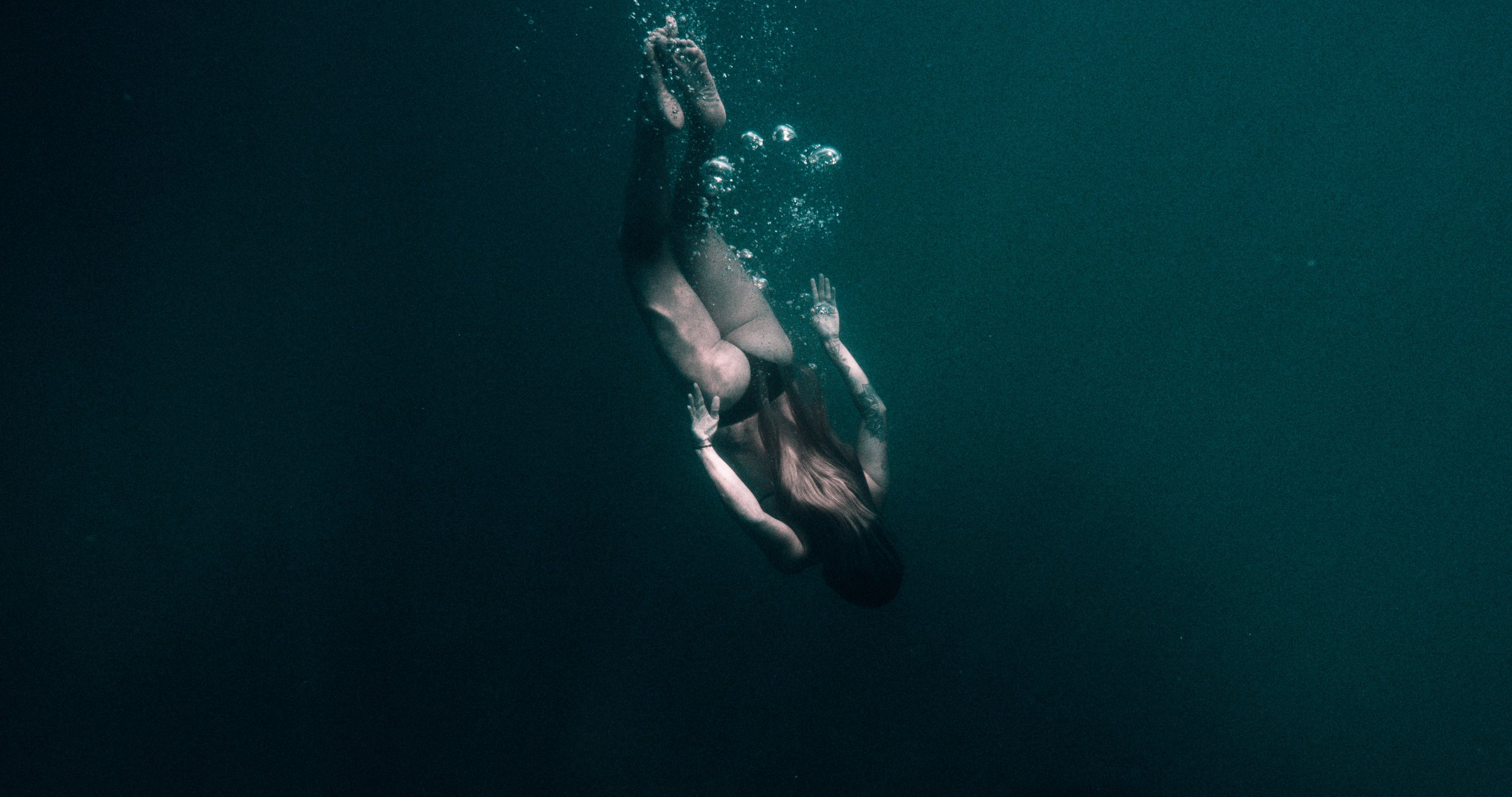 Underwater shot of a woman diving down into dark teal water with sunlight and bubbles. The woman has long hair and tattoos.