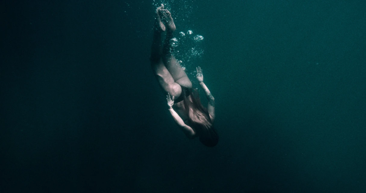 Underwater shot of a woman diving down into dark teal water with sunlight and bubbles. The woman has long hair and tattoos.