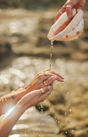Close-up of hands holding a large conch shell pouring water over cupped hands adorned with gold rings against a rocky background