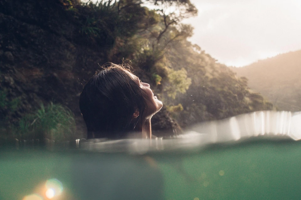 Adult female in water with lush greenery behind; face tilted up, enjoying nature. Aquatic scene with foliage, sky, and natural lighting.