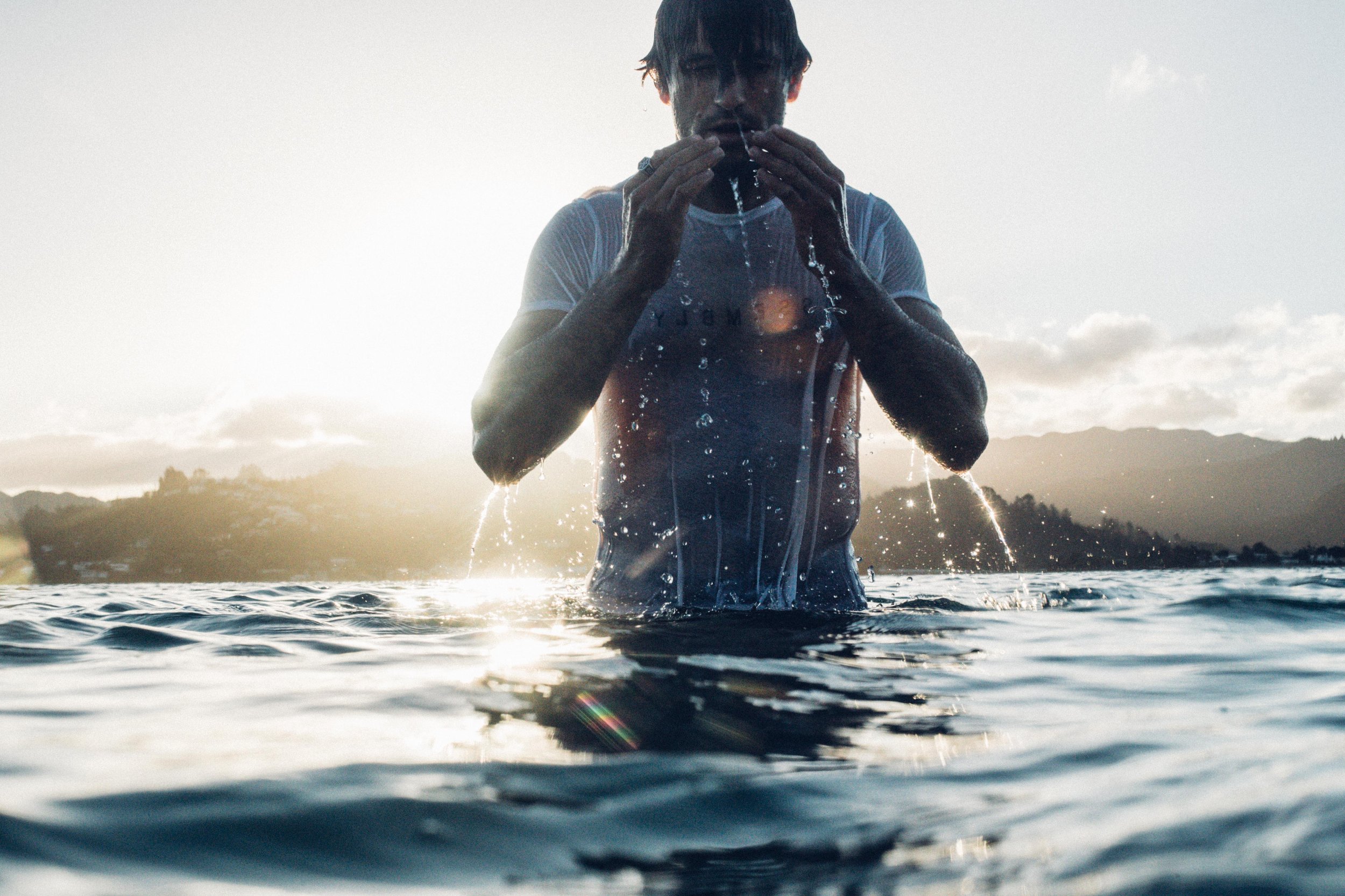 Man emerges from the ocean, wearing a wet white t-shirt, with water dripping. He is surrounded by water, with mountains in the background.