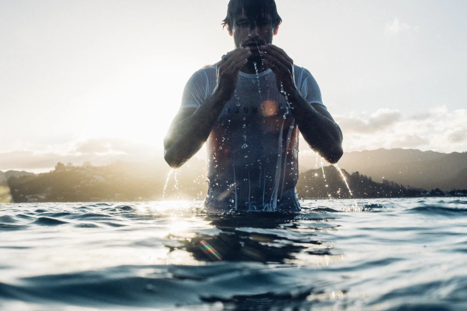 Man emerges from the ocean, wearing a wet white t-shirt, with water dripping. He is surrounded by water, with mountains in the background.