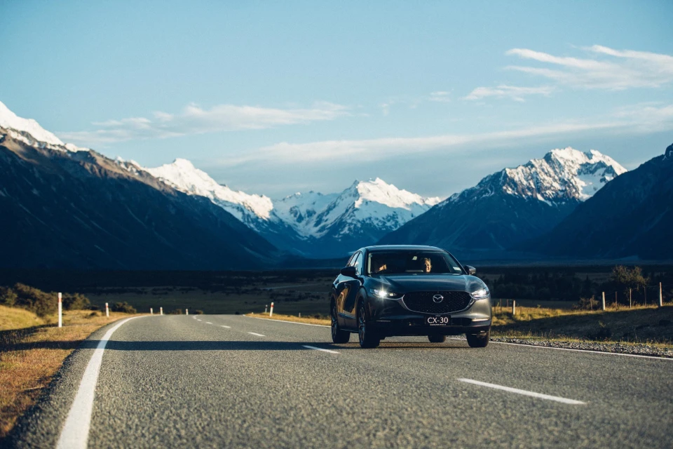 Mazda CX-30 driving on an open road with snow-covered mountains in the background under a clear blue sky, showcasing automotive travel.
