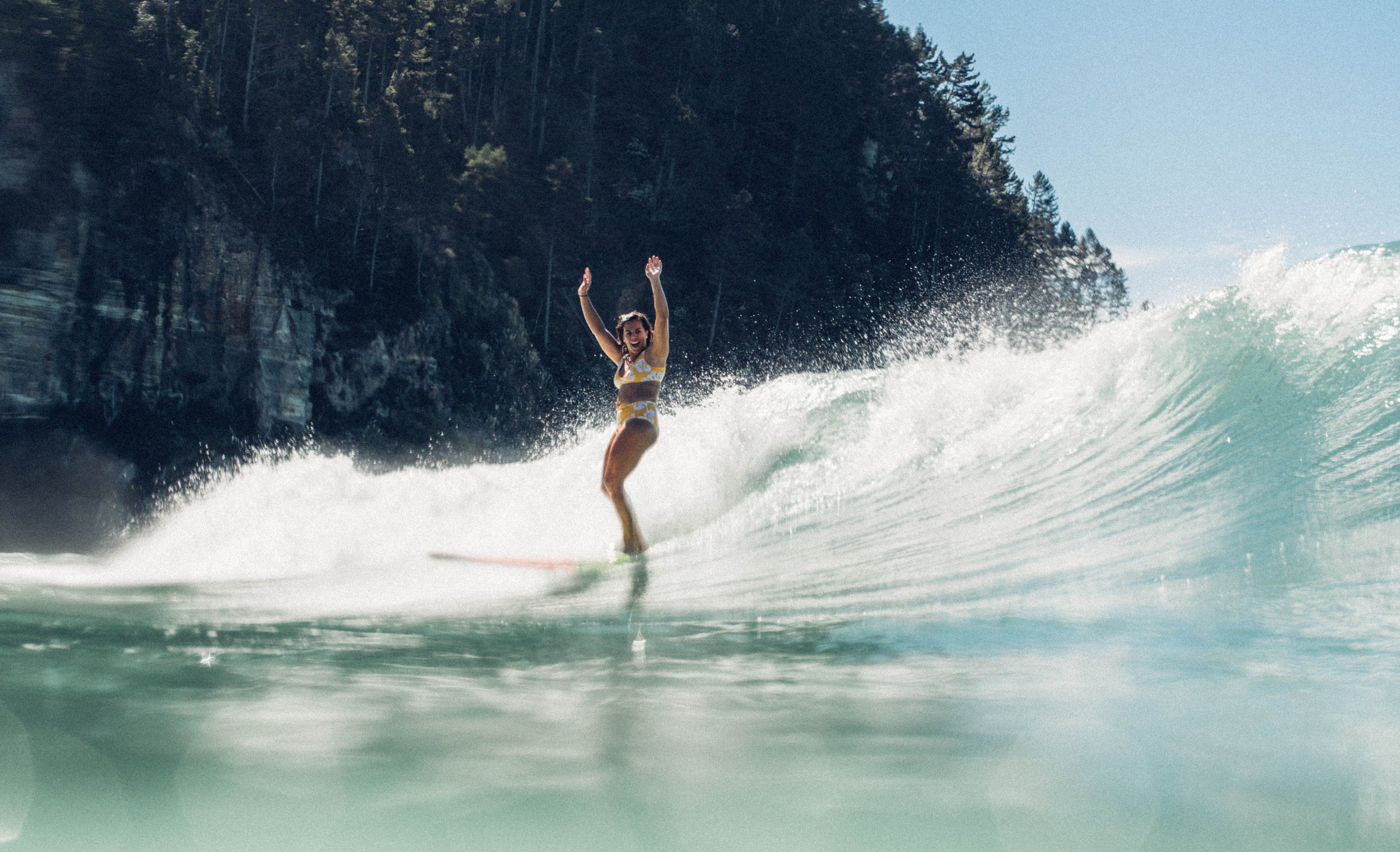 Woman surfs a wave near a rocky coastline, captured from an underwater perspective. She wears a bikini and raises her arms, enjoying the sport.