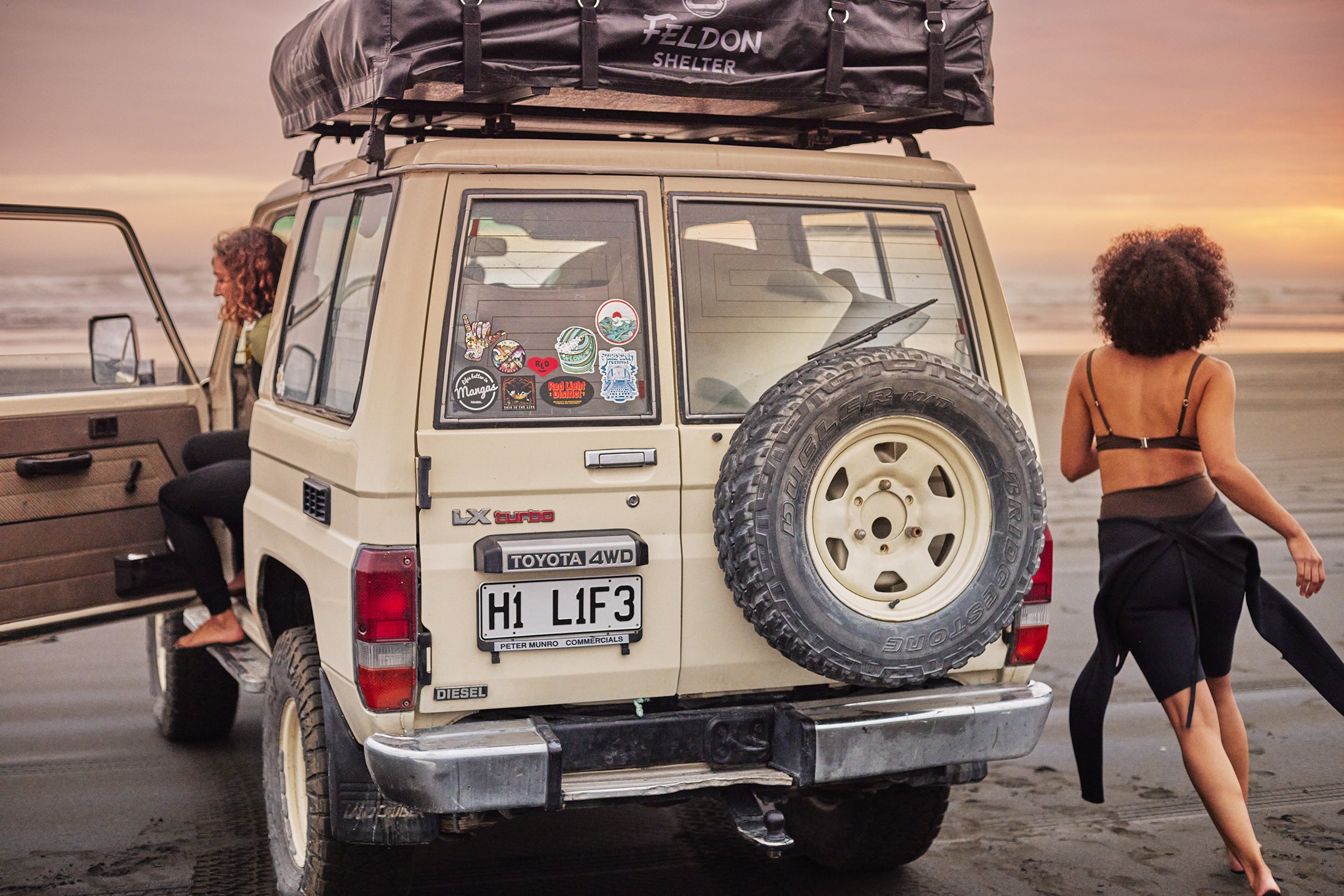 Tan four-wheel drive vehicle on a beach at sunset with two women, one with a surfboard, enjoying an outdoor adventure.