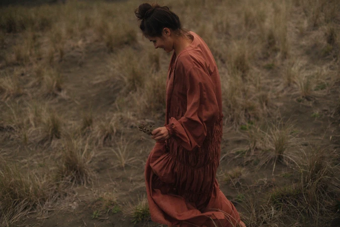Woman with dark hair in a topknot wearing a terracotta dress walking through a dry grassy field