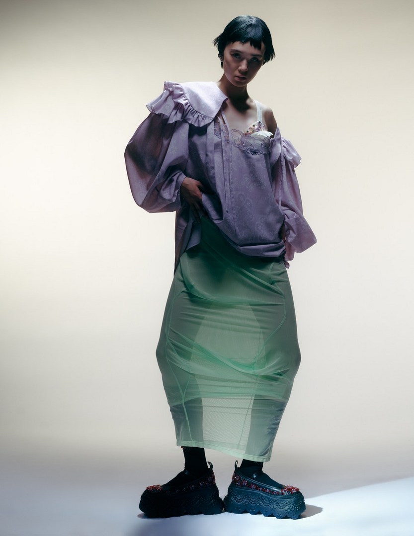 A young adult fashion model poses in an oversized lavender shirt with a white collar, sheer green skirt, and chunky black floral shoes.