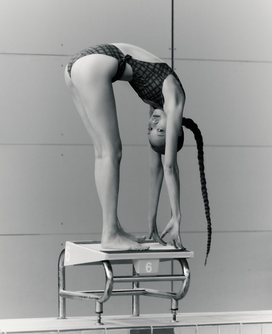 Monochromatic image of a female athlete poised on a diving board, arching her back before diving into a swimming pool.