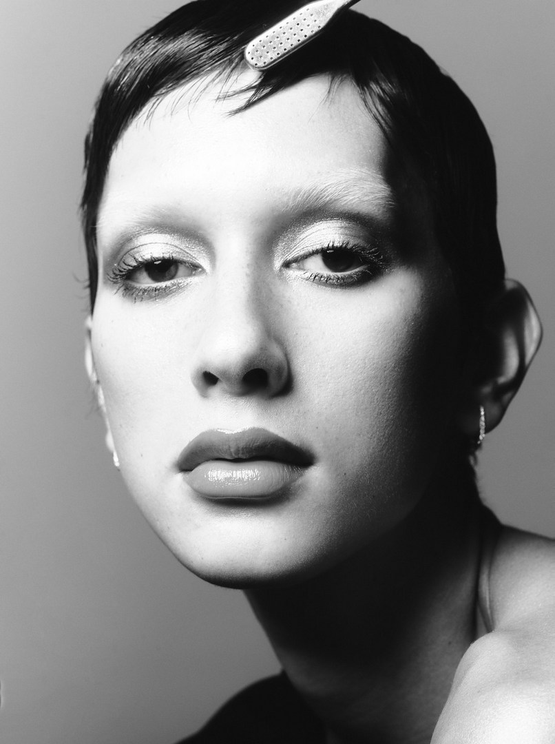 Monochrome portrait of a young adult woman with short hair and neutral expression, wearing makeup, and a hair clip, against a plain grey background.