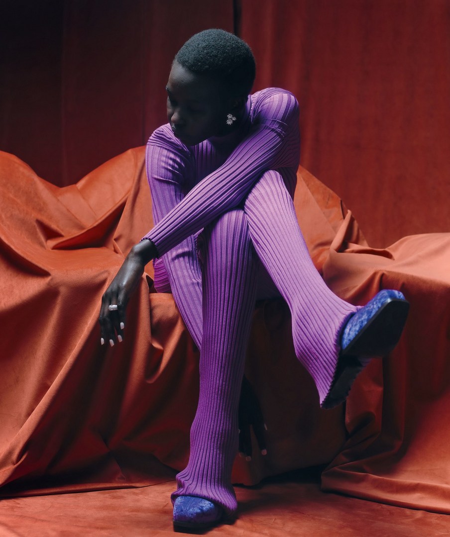 A slender Black woman models purple ribbed knitwear with blue crushed velvet shoes, seated against draped reddish-orange textile backdrop.