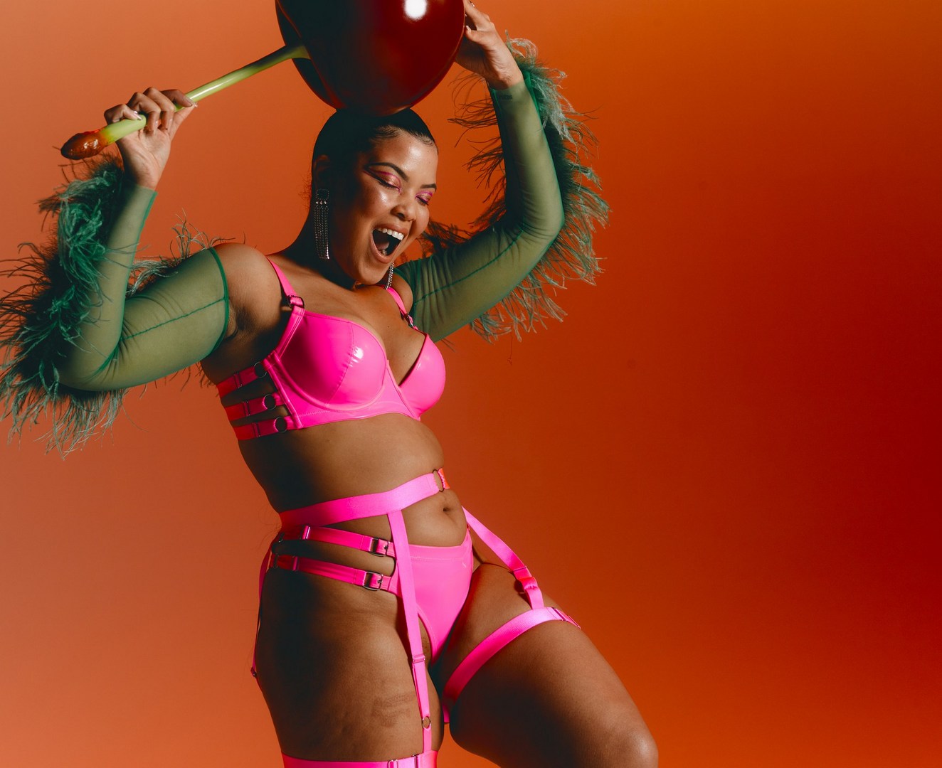 An adult female model strikes a playful pose in a neon pink lingerie set, set against a vibrant orange gradient backdrop in a studio.