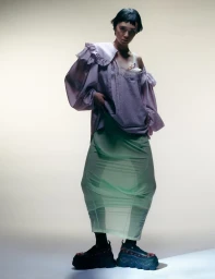 Model wearing a ruffled lavender top and sheer green skirt against a plain studio background.
