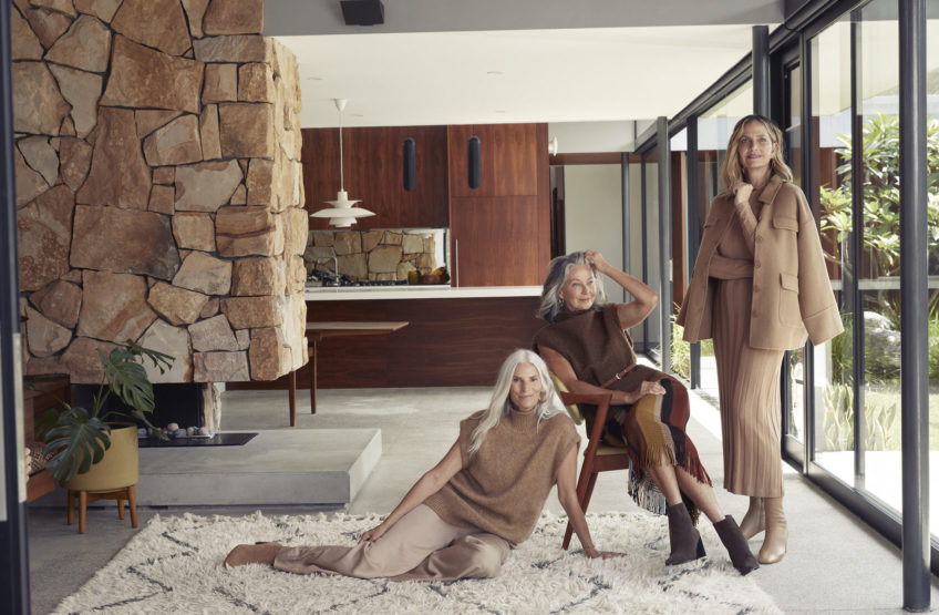 Three adult women in a modern home interior with stone fireplace and large windows. The women are wearing casual earth-toned clothing.