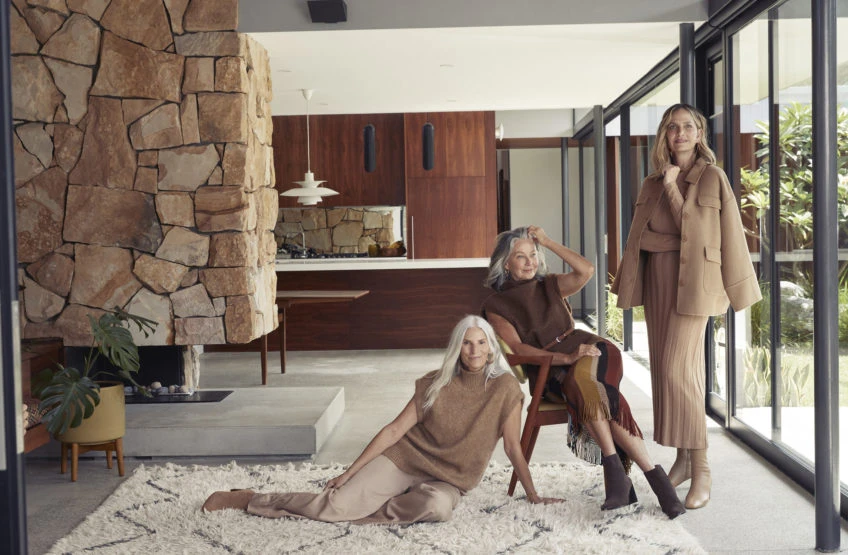 Three adult women in a modern home interior with stone fireplace and large windows. The women are wearing casual earth-toned clothing.