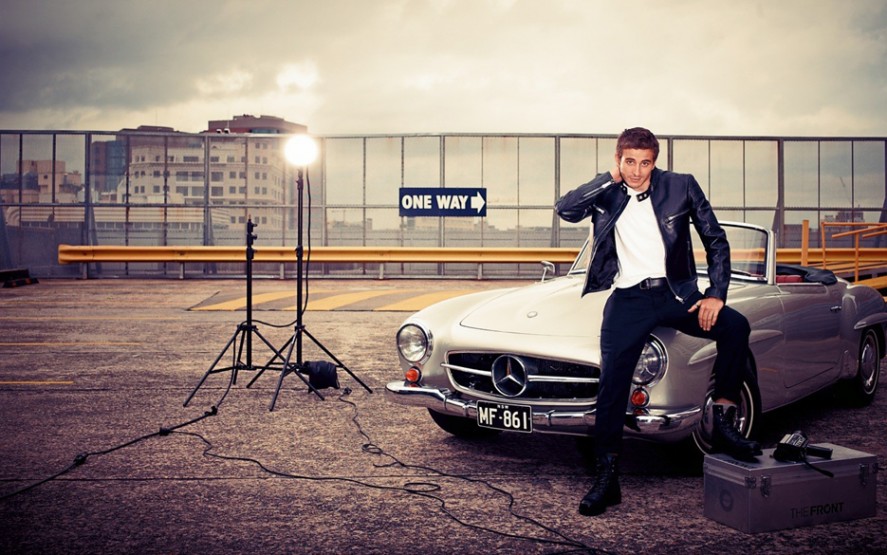 Stylish adult male posing with a silver convertible car, illuminated by studio lighting, set on a rooftop parking lot.