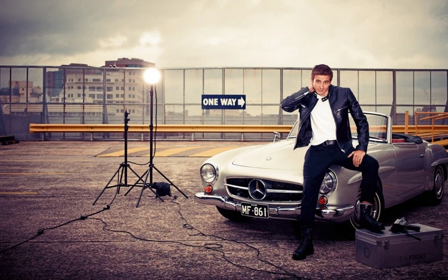 Stylish adult male posing with a silver convertible car, illuminated by studio lighting, set on a rooftop parking lot.