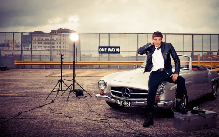 Stylish adult male posing with a silver convertible car, illuminated by studio lighting, set on a rooftop parking lot.