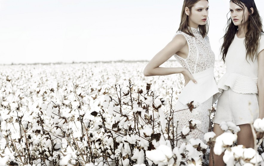 Two fashion models in white outfits pose in a cotton field under bright daylight, showcasing minimalist styles and textured materials.