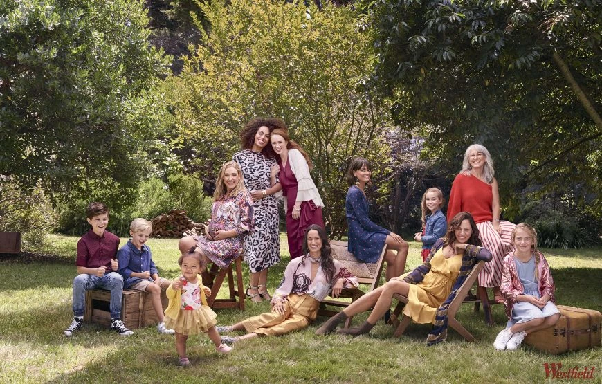 A diverse group of women and children are gathered outdoors, sitting and posing on wooden crates, chairs, and a vintage suitcase in a yard with trees.