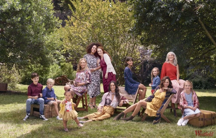 A diverse group of women and children are gathered outdoors, sitting and posing on wooden crates, chairs, and a vintage suitcase in a yard with trees.