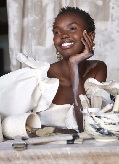Smiling Black woman poses in a white dress with large ruffles, holding her head, with paint brushes, bowls, and neutral drapes in background.