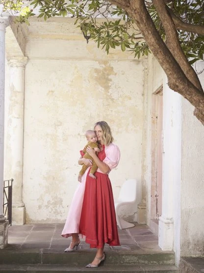 A fair-skinned woman in a red and pink dress and heels holds a baby on the steps of a building with stone columns and foliage.