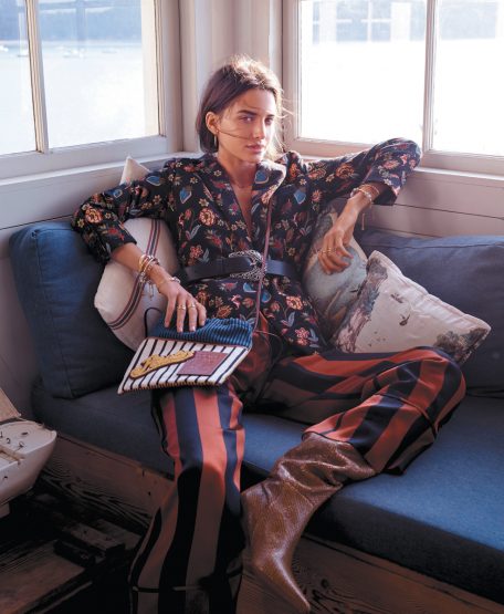 Woman in patterned blazer and striped pants sits indoors with a patterned purse. Natural window light illuminates the scene.