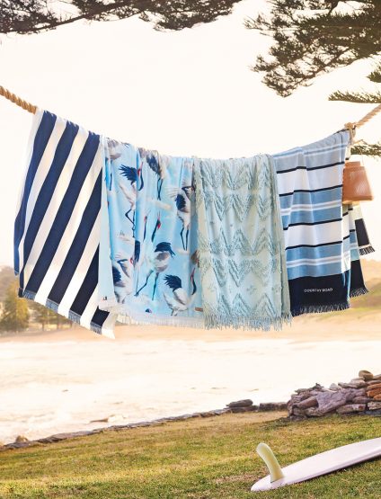 Beach towels with various patterns hang from a rope outdoors in front of a grassy area near a beach and the ocean in a serene setting.