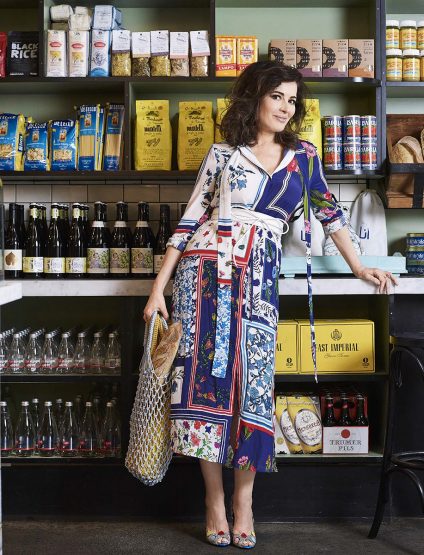 Woman in a floral dress stands in front of shelves of food products, holding a mesh bag with baguettes, creating a lifestyle scene.