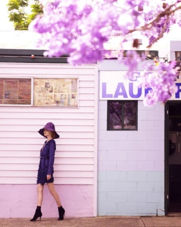 Model in purple hat and blue dress walking past pastel buildings framed by purple flowers