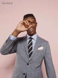 A smiling black man in a grey plaid suit holding his hand near his face against a solid pink studio backdrop.
