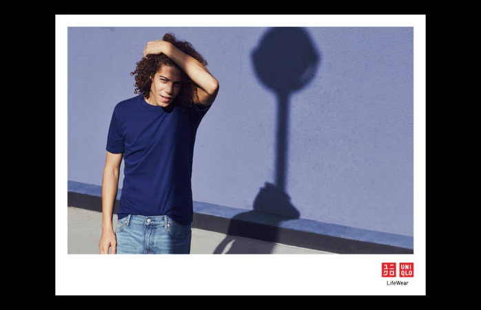 Young man with curly brown hair wearing a blue t-shirt and blue jeans posing in front of a purple textured wall.