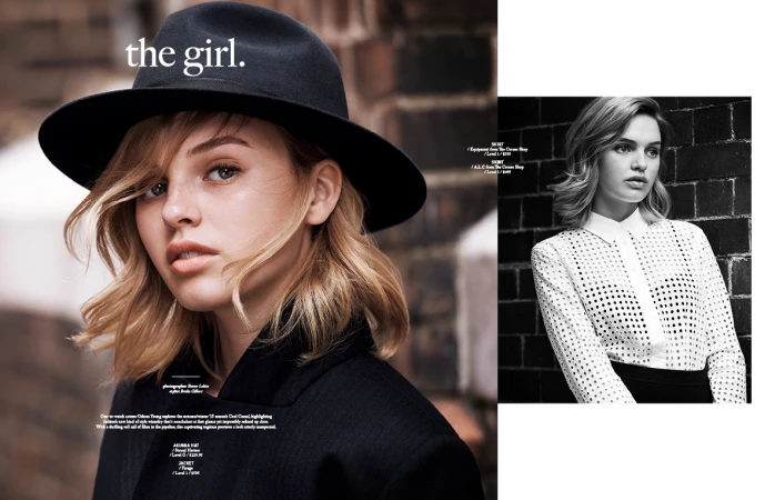 Close-up of a young woman with blonde hair wearing a black fedora hat and dark coat against a brick wall.