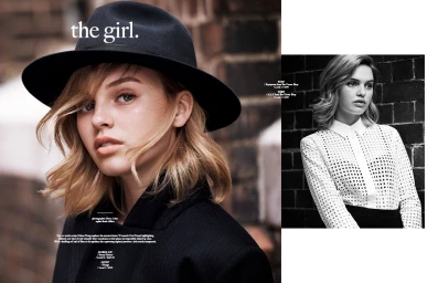 Close-up of a young woman with blonde hair wearing a black fedora hat and dark coat against a brick wall.