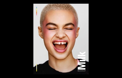Close up portrait of a person with short blonde buzzcut hair wearing pink makeup laughing against a plain white background